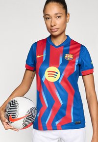 Μπλούζα γυναικείου FC Barcelona με κόκκινο και μπλε ζιγκζαγκ μοτίβο, στρογγυλή λαιμόκοψη, κίτρινο λογότυπο Nike και λεπτομέρεια κυκλικού λογότυπου Spotify, συνοδευόμενη από μαύρη και λευκή μπάλα.