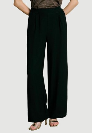 Pantalon noir taille haute à jambes larges porté avec des sandales à talons dorées à brides et un haut métallique foncé, les mains dans les poches.