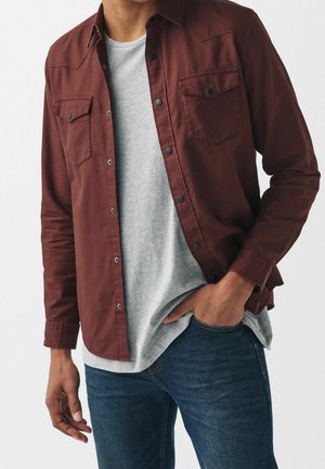 Mand iført uopknappet mørkerød skjorte over lysegrå t-shirt med den ene hånd i lommen på blå jeans mod hvid baggrund.