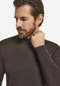 Ciemnobrązowy golf o gładkiej fakturze, dopasowany fason z długimi rękawami. Kołnierz lekko podwinięty, dłoń trzymająca kołnierz.