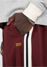 Burgundy tepláky so bočnými vreckami, s bielymi a námorníckymi pruhmi a zlatou vyšívanou aplikáciou na ľavej strane. Hladký textilný povrch.