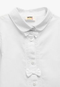 Camicia bianca in cotone per bambini con colletto, chiusura frontale a cinque bottoni, dettagli in pizzo e due piccoli fiocchi in tessuto sul petto.