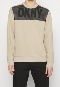 Felpa beige con pannello in rete nera caratterizzato dal logo "DKNY", maniche lunghe, polsini e orlo a costine, texture liscia, vestibilità casual.