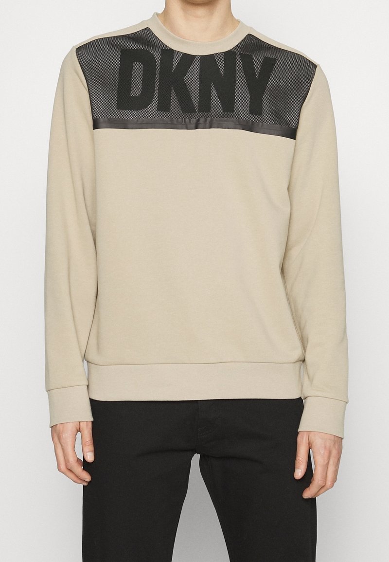 Felpa beige con pannello in rete nera caratterizzato dal logo "DKNY", maniche lunghe, polsini e orlo a costine, texture liscia, vestibilità casual.