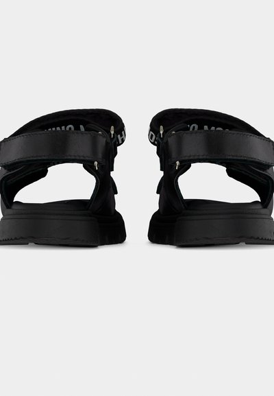Ζευγάρι μαύρα σανδάλια με χοντρές σόλες και ρυθμιζόμενους πίσω ιμάντες με λευκό τυπωμένο κείμενο, εμφανιζόμενα από πίσω σε λευκό φόντο.