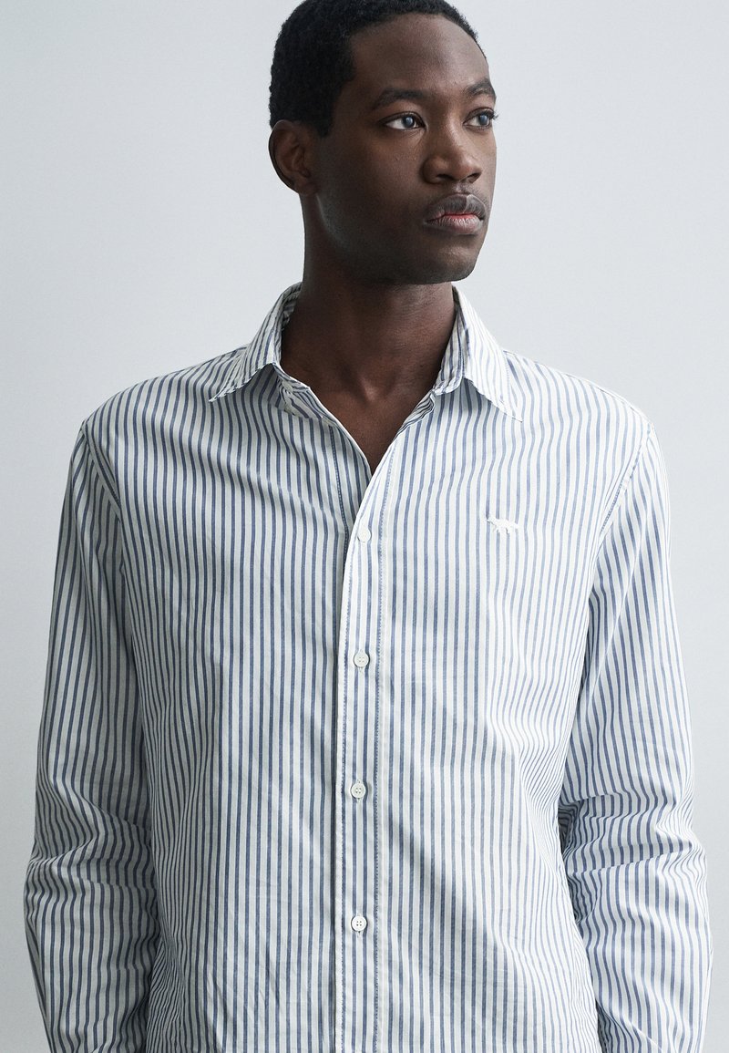 Riba punaste ja valgete triipudega pikkade varrukatega särk helesinisest puuvillast. Omab nuppe avanevat kaelust ja väikest tikandiga logo rinnal.