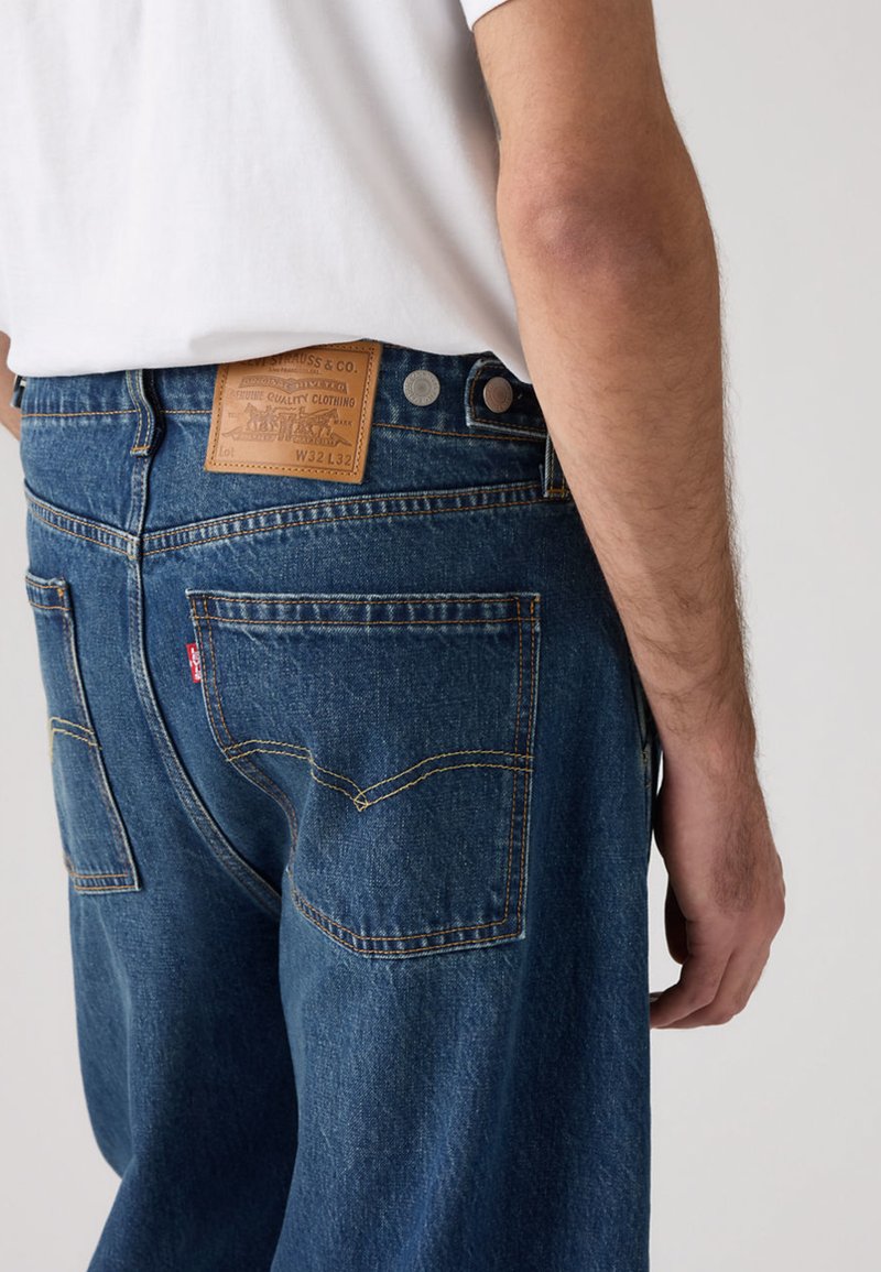 Vaqueros de mezclilla de color azul medio, con una etiqueta de cuero en la parte trasera, herrajes de cobre y costuras contrastantes en los bolsillos.
