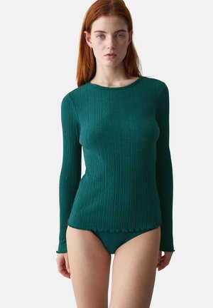 Vrouw met rood haar die een aansluitend donkergroen geribbeld topje met lange mouwen en bijpassend hoog uitgesneden ondergoed draagt, staande tegen een effen achtergrond.
