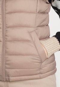 Prošívaná vesta v světle taupe barvě s vertikálním prošitím, zapínáním na zip a bočními kapsami. Žebrovaný manžet v béžové barvě vykukuje zpod rukávu.