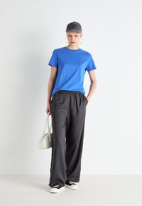Camiseta de algodón azul, pantalones de punto gris sueltos, zapatillas negras y una gorra gris. El modelo sostiene un pequeño bolso blanco. Un atuendo simple y casual.