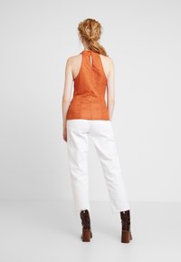 En rostorange ärmlös topp med hög hals, strukturerat mönster och detaljer med nyckelhål bak, kombinerad med vita cropped byxor och bruna klackstövlar.