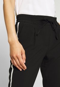 Svarta joggingsbyxor med elastisk midja och snörning, sidofickor och ett vitt rand längs yttersemsorna. Slät tygtextur.