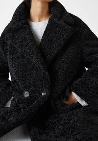 Manteau noir et duveteux avec un large col, un design croisé et de gros boutons, présentant un tissu texturé et des poches profondes.