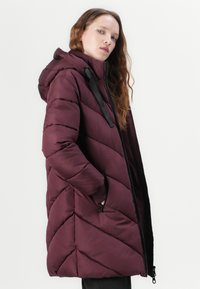 Burgunderfarget pufferjakke med chevron-quilting, hette, glidelåser og svarte detaljer. Laget av lett materiale, med en avslappet passform.