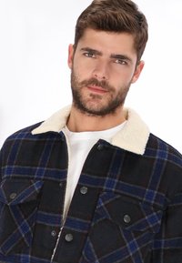 Chaqueta de franela para hombre con un patrón de cuadros en negro y azul, cuello de piel sintética blanca, botones de presión y dos bolsillos en el pecho.