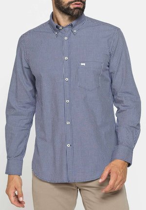 Uomo che indossa una camicia a quadri blu navy e bianca con bottoni, maniche lunghe e pantaloni beige su sfondo bianco.