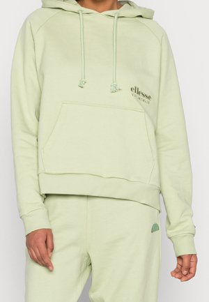 Persoon draagt een lichtgroene ellesse-hoodie en bijpassende joggingbroek, voorkant zichtbaar, met kangoeroezak en merklussen op borst en dij.