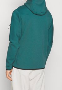 Person som bär en turkos huvtröja med svarta detaljer och vita byxor, sedd bakifrån mot en enfärgad bakgrund.