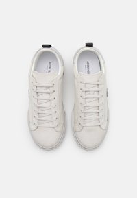 Baskets en daim blanc avec lacets plats, bout rond et une languette noire au talon. Elles présentent un patch logo sur le côté.
