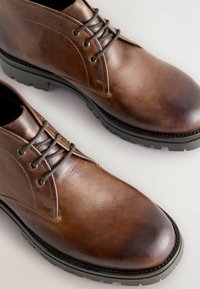 Botine din piele maro cu o finisare netedă, având vârfuri rotunde, ochiuri metalice și șireturi închise la culoare. Tălpile groase din cauciuc oferă aderență.