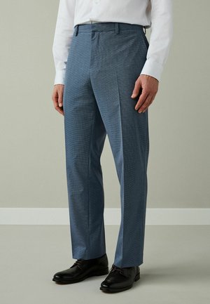 Mann in schlanken blauen Hosen, schwarzen Anzugschuhen und weißem Langarmhemd, steht drinnen auf einem hellgrauen Boden.