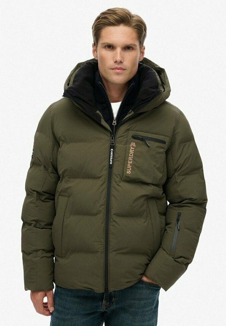 Młody mężczyzna w oliwkowej puchowej kurtce Superdry z zamykanymi kieszeniami i kapturem, stojący na tle białego tła.