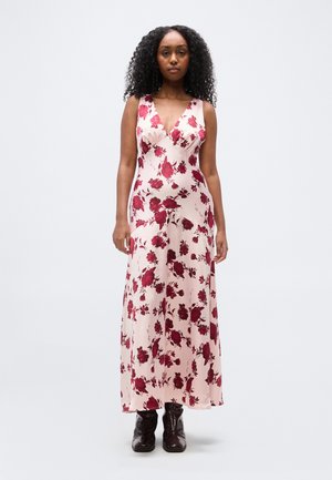 Femeie cu păr creț purtând o rochie fără mâneci, roz pal, cu imprimeu floral roșu și cizme înalte închise la culoare, stând pe un fundal simplu.