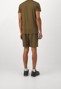 Sada sportovního trička a kraťasů olivově zelené barvy, vyrobená z lehké látky, s černým logem na rukávu, nošená s černými sportovními botami.