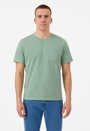 Grünes kurzärmliges T-Shirt aus Baumwolle mit rundem Ausschnitt und einer Brusttasche. Das Modell trägt blaue Jeans.