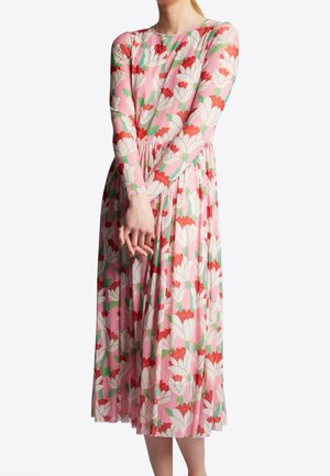 Femme portant une robe midi rose à manches longues avec un grand imprimé floral blanc et rouge, les mains jointes devant, debout sur fond blanc.