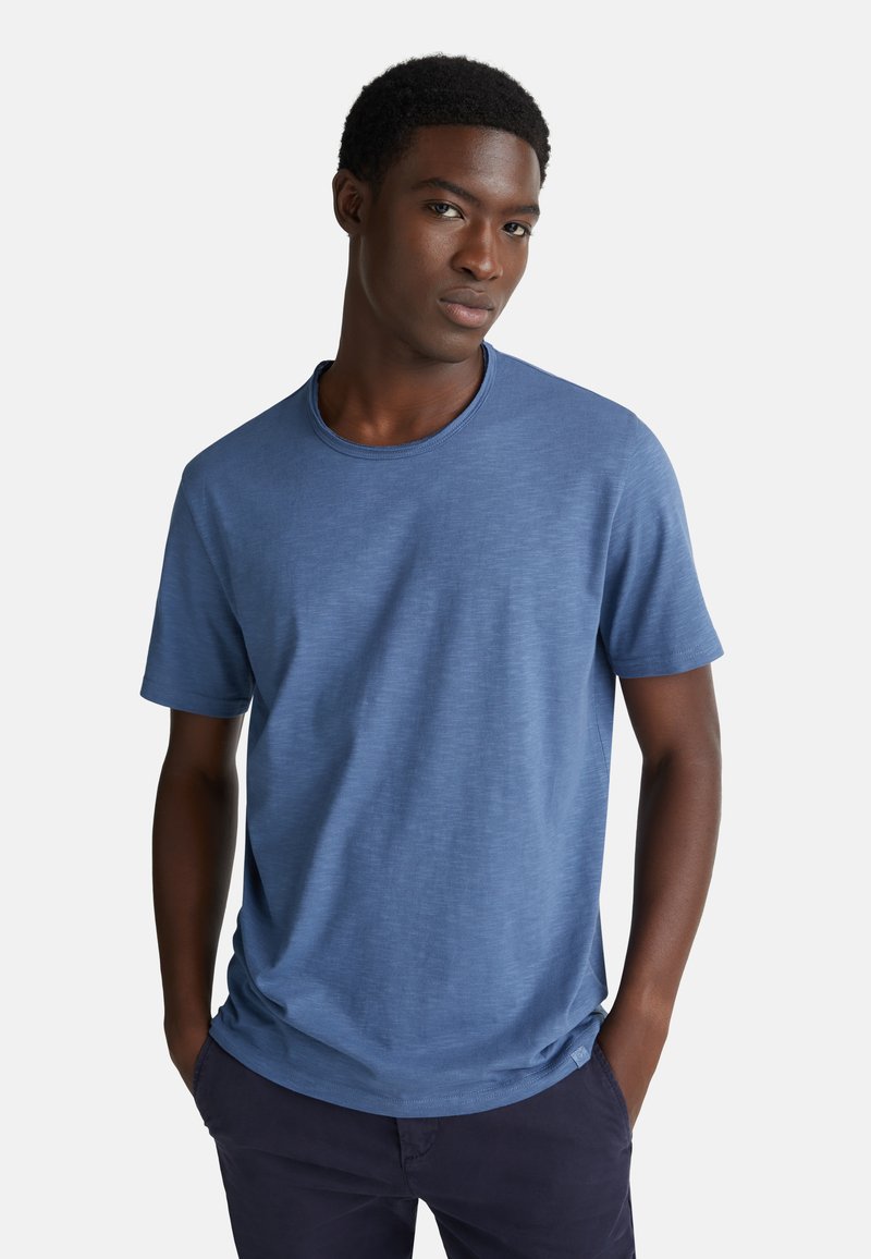 Mladý muž s krátkými kudrnatými vlasy, oblečený v modrém tričku s krátkým rukávem a tmavých kalhotách, se zdrženlivě kouká s rukama v kapsách na světlém pozadí.