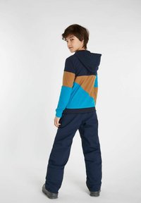 Bunte Kapuzenjacke mit blauen, marineblauen und braunen Paneelen; weicher Stoff; kombiniert mit dunkelblauen Hosen und schwarzen Stiefeln; lässige Passform.