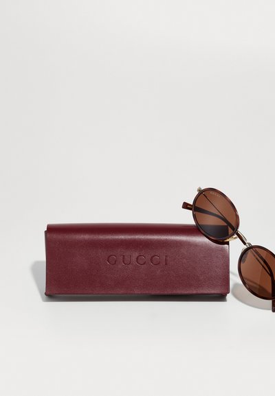 Gucci aurinkolasit, joissa on ruskeat linssit ja pyöreä muotoilu, lepäävät viininpunaisen nahkakuoren vierellä, johon on painettu brändin nimi.