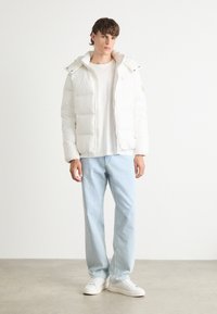 Vit puffjacka med huva, vadderad textur, dragkedja fram och två sidofickor. Bärs över en ljus skjorta och kombineras med ljusblå jeans och sneakers.