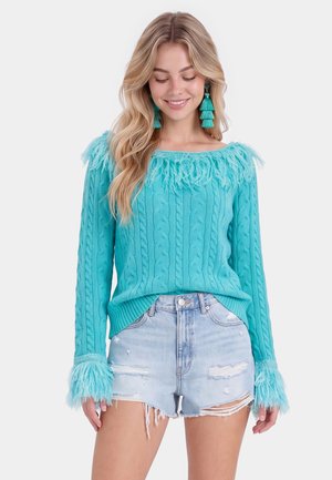 Kvinde iført en turkis kabelstrik sweater med frynser på kraven og ærmekanter, kombineret med lyse, revnede denimshorts, der smiler med lukkede øjne.