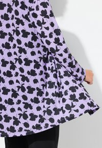 Violetti kangasblouse, jossa on musta kukkakuosi, virtaava muotoilu ja pitkät hihat. Helma on hieman levenevä.