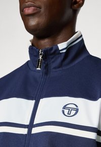 Marineblauwe zip-upjack met een hoge kraag, voorzien van witte en lichtblauwe strepen over de borst en een cirkelvormig logo aan de linkerkant.