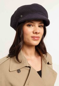 Bonnet en laine foncée avec un large bord, arborant un accent logo métallique. Présenté sur un modèle portant un manteau beige.