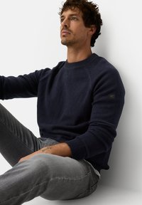Navy blue knitted sweater with ribbed cuffs and neckline, worn with gray slim-fit jeans. Features a small logo on the left sleeve.