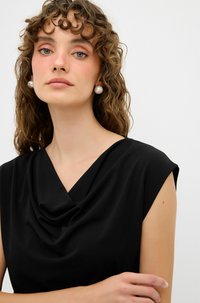Černý top bez rukávů s volným výstřihem. Hladká tkanina a uvolněný střih. Modelka má na sobě velké perlové náušnice.