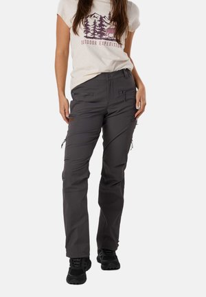 Frau trägt beige T-Shirt mit Outdoor-Grafik und graue Wanderhose mit Reißverschlusstaschen und schwarzen Wanderschuhen, steht vor weißem Hintergrund.