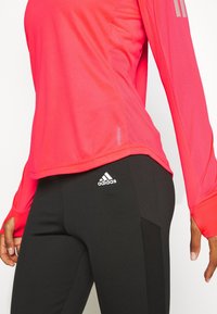Top sportivo a maniche lunghe di colore rosa acceso con dettagli in mesh, abbinato a leggings neri con logo Adidas bianco. Texture liscia, design aderente.