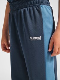 Kind trägt blaue Sporthose mit elastischem Bund und „hummel performance“-Logo am oberen Oberschenkel.