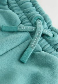 Detailný záber na tyrkysovú textíliu sťahovanú šnúrkou, zdobenú štruktúrovanými vzormi a štítkom s nápisom „BY TED BAKER“ v kontrastnom písme.