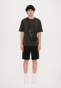 Zwart gestreept T-shirt met geometrische ontwerpen, zwarte shorts, een kleine zwarte schoudertas en witte sneakers met zichtbare veters.