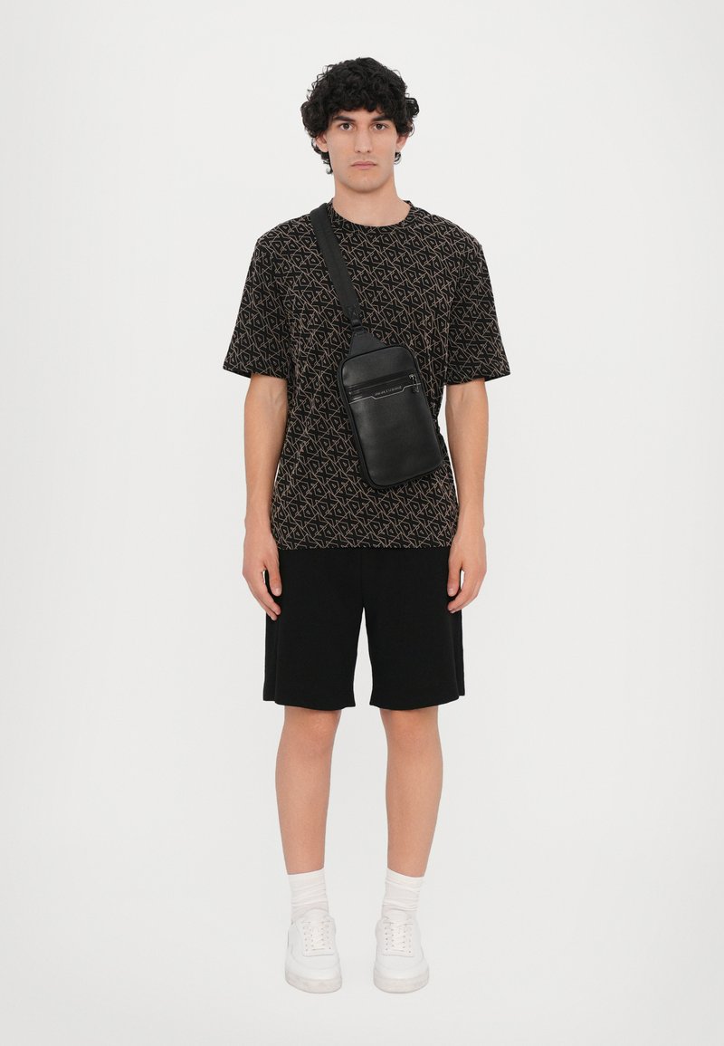 Zwart gestreept T-shirt met geometrische ontwerpen, zwarte shorts, een kleine zwarte schoudertas en witte sneakers met zichtbare veters.
