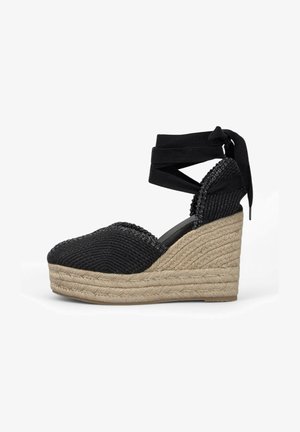 Zwarte wedge espadrille met geweven textuur, enkelbandjes en een jute platform. Heeft een ronde neus en decoratieve stiksels langs de bovenkant.