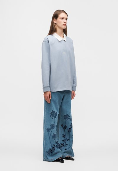 Ανοιχτό μπλε πουκάμισο μακρυμάνικο με λευκό γιακά και μικρή ετικέτα· παντελόνι τζιν με φαρδιά πόδια και σκούρα κεντήματα με λουλούδια· μαύρα μυτερά παπούτσια.