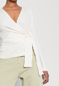 Top bianco avvolto con scollatura a V, tessuto texturizzato, maniche lunghe e vita legata. Indossato con pantaloni verde chiaro, che mostrano uno stile casual.