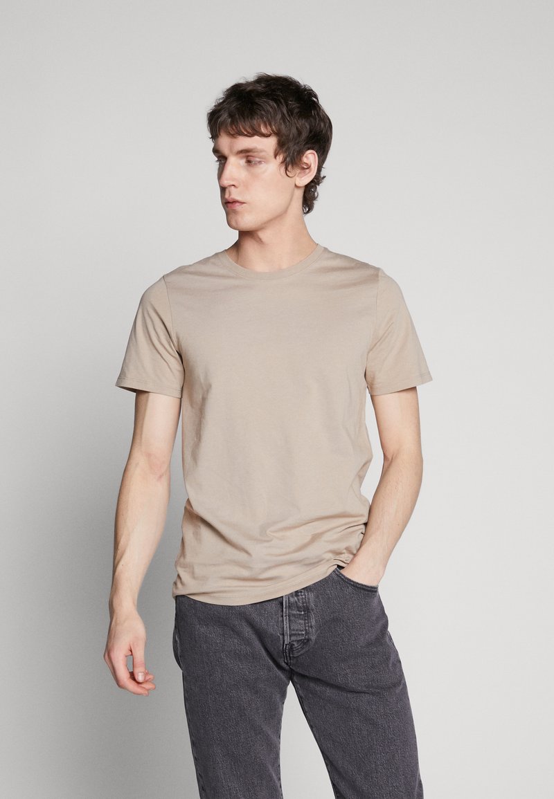 Light beige short-sleeve t-shirt, round neckline, casual fit, smooth texture. Paired with dark gray jeans, hands in pockets.